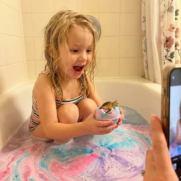 Child in colorful bath water holding a bath bomb with a toy turtle; photo taken on a phone